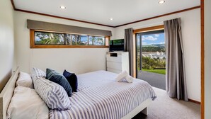 Een slaapkamer met een bed, kussens, een televisie, een bureau en uitzicht op groen door een raam.