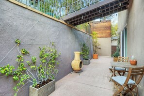 Eine Terrasse im Freien mit Betonboden, Holzstühlen, einem Tisch, Topfpflanzen und einer Pergola mit hängenden Lichtern.