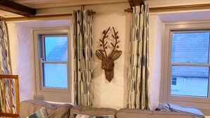 A living room with a sofa, patterned curtains, a wall-mounted deer head decoration, and windows with a view of the outside. - Spacious Seaside Cottage in Dale, Pembrokeshire – Steps from the Beach, Sleeps 9 (Haverfordwest)