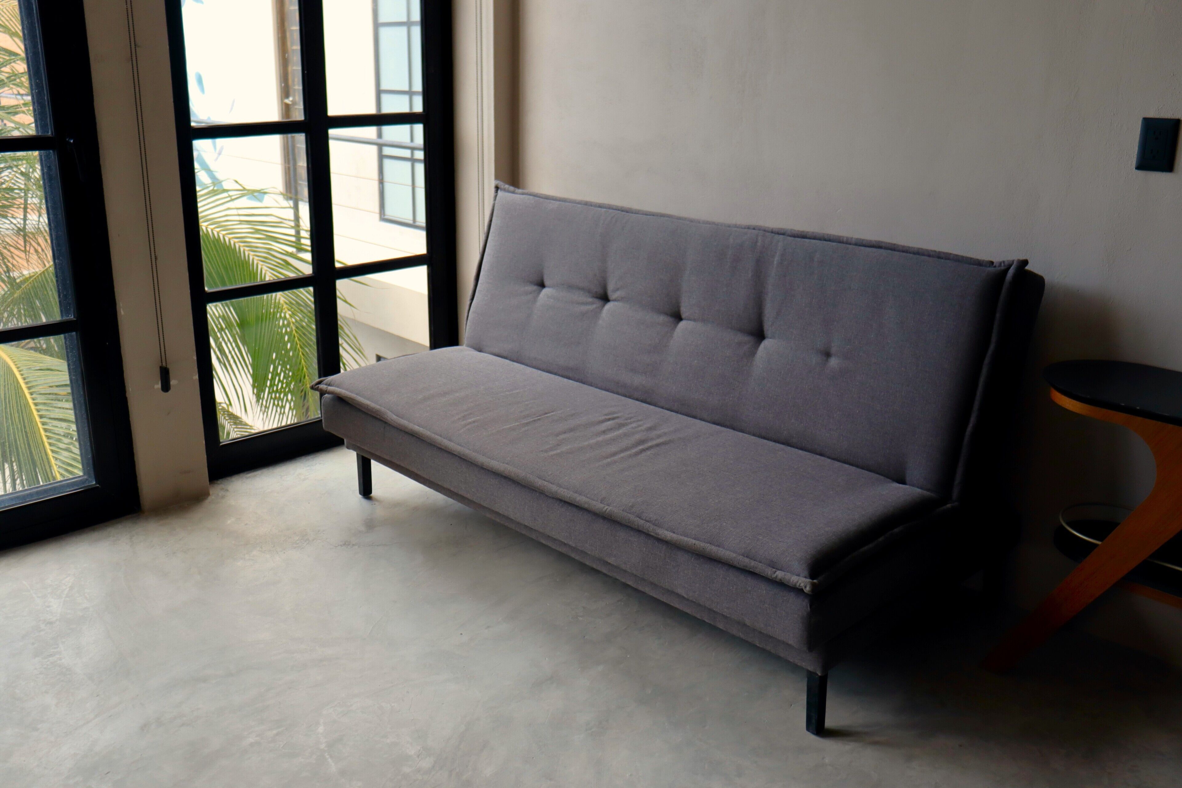 Un sofá gris en una habitación con piso de concreto y una ventana con vista a palmeras.