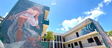 Um edifício com um grande mural de uma mulher e peixes na lateral, vários andares e um céu azul claro.