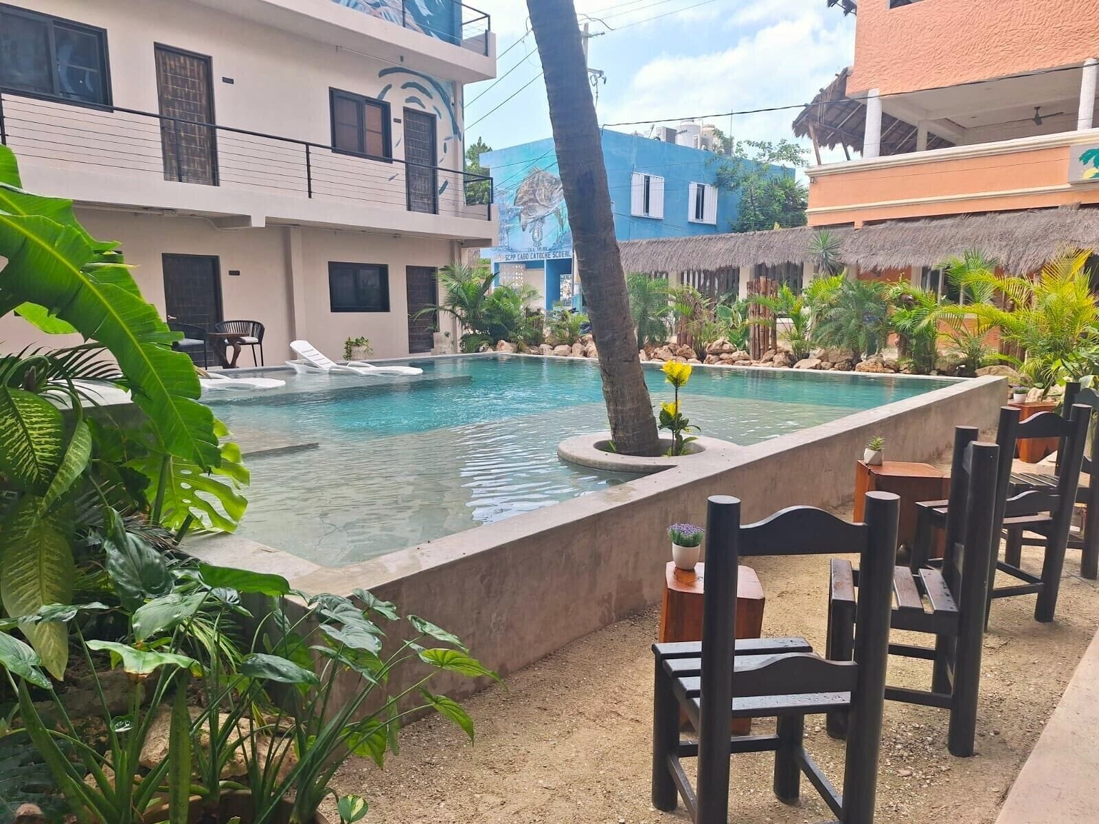 Una piscina con sillas de descanso, rodeada de plantas tropicales y un edificio con balcones.
