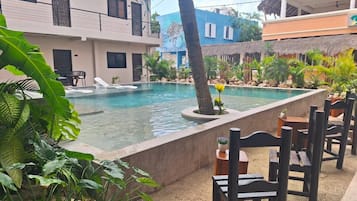 Uma piscina com espreguiçadeiras, rodeada por plantas tropicais e um edifício com varandas.