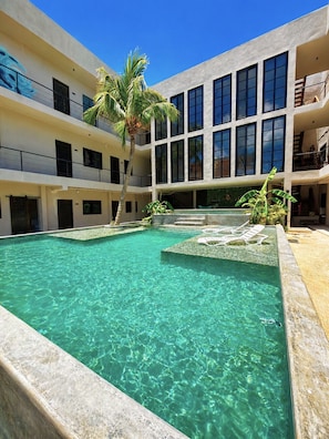 Un edificio de varios niveles con piscina central, palmeras y cielo azul despejado.