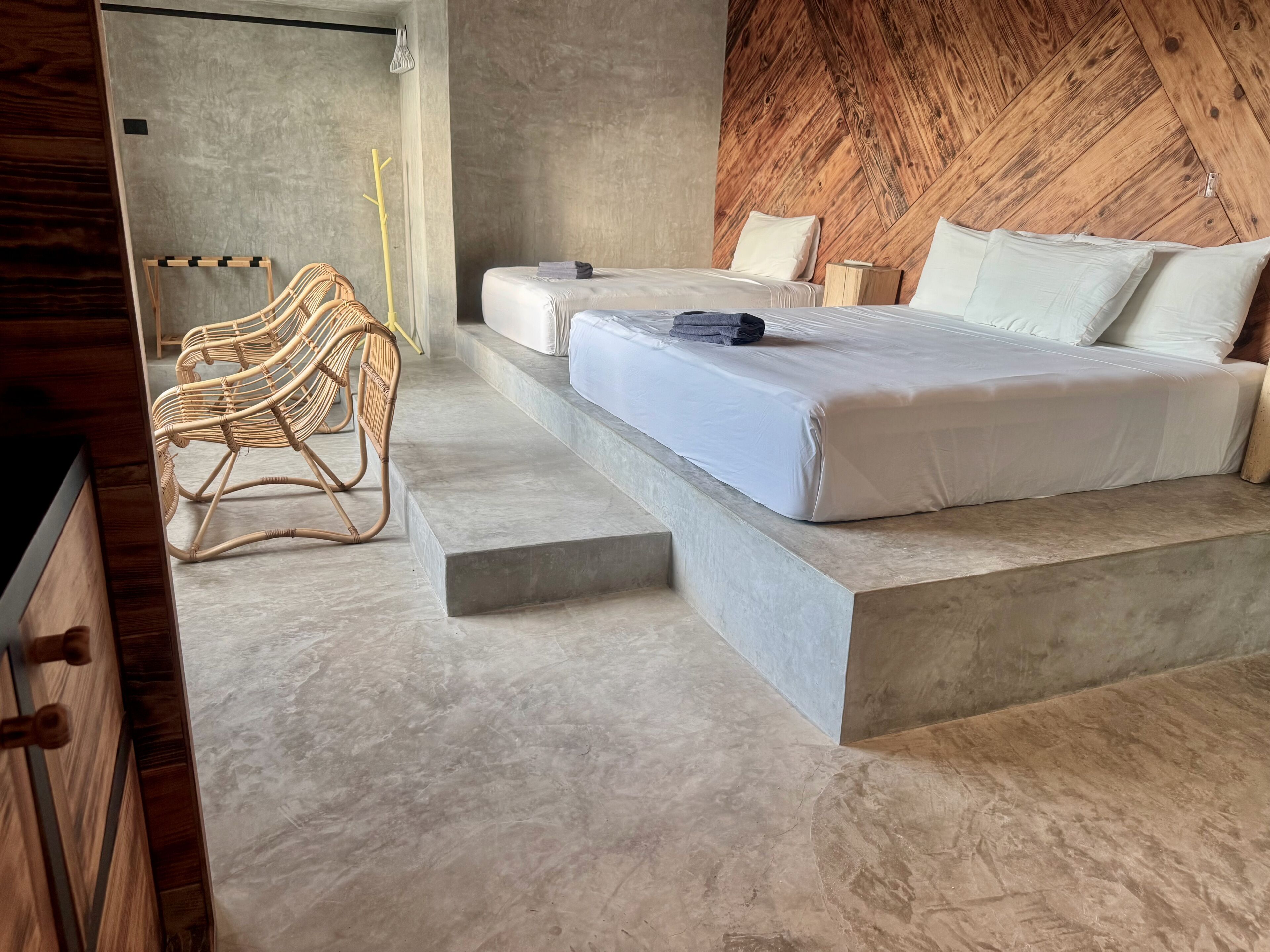 Un dormitorio moderno con una cama, dos mesitas de noche, una silla de mimbre y una plataforma de concreto.