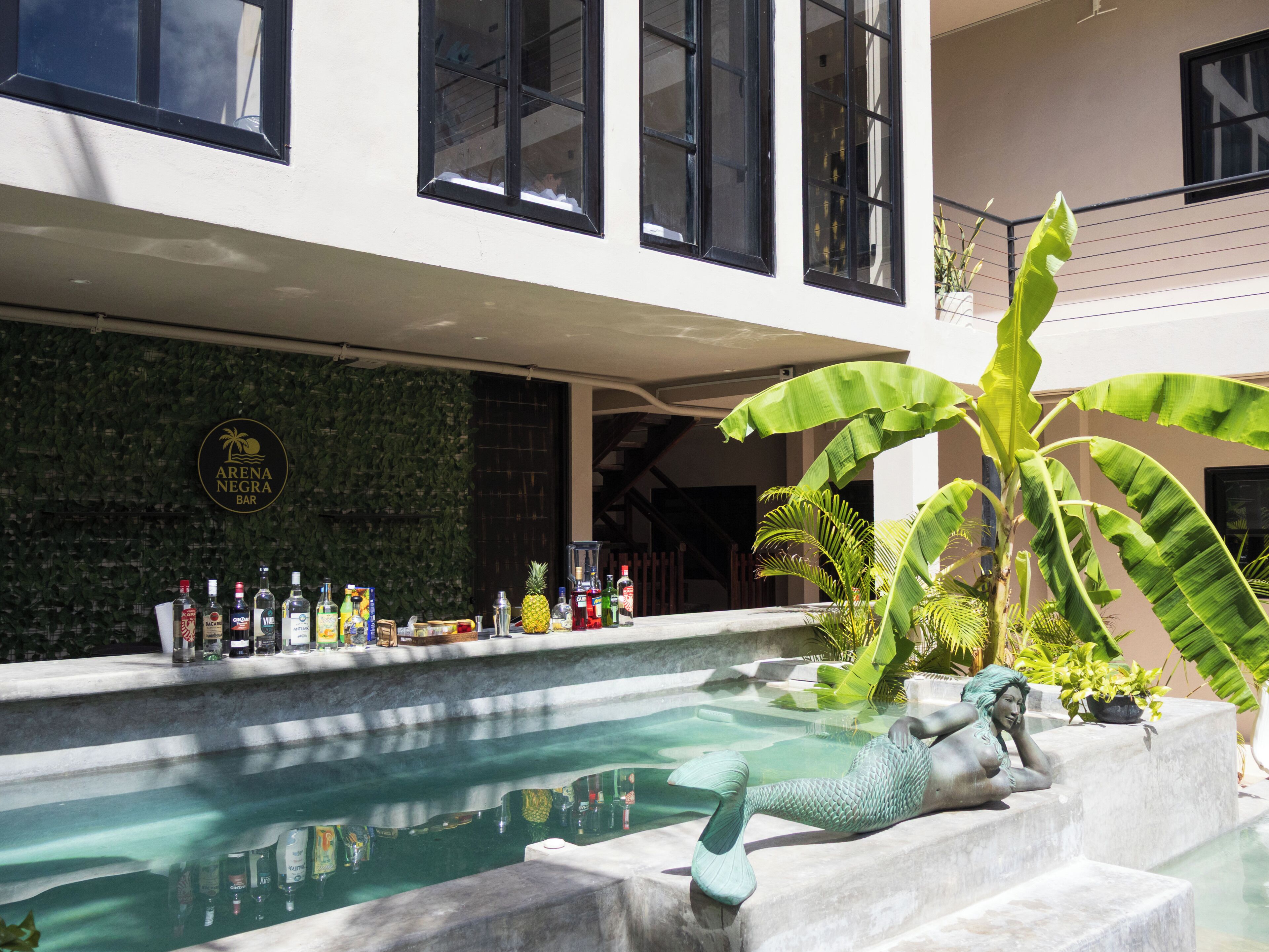 Zona de piscina con una estatua, un bar con diferentes botellas y una pared con plantas y un letrero.