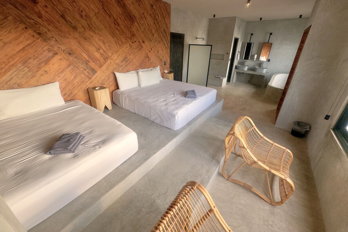 A modern hotel room with two beds, a wooden accent wall, a sitting area with wicker chairs, and a bathroom visible in the background.