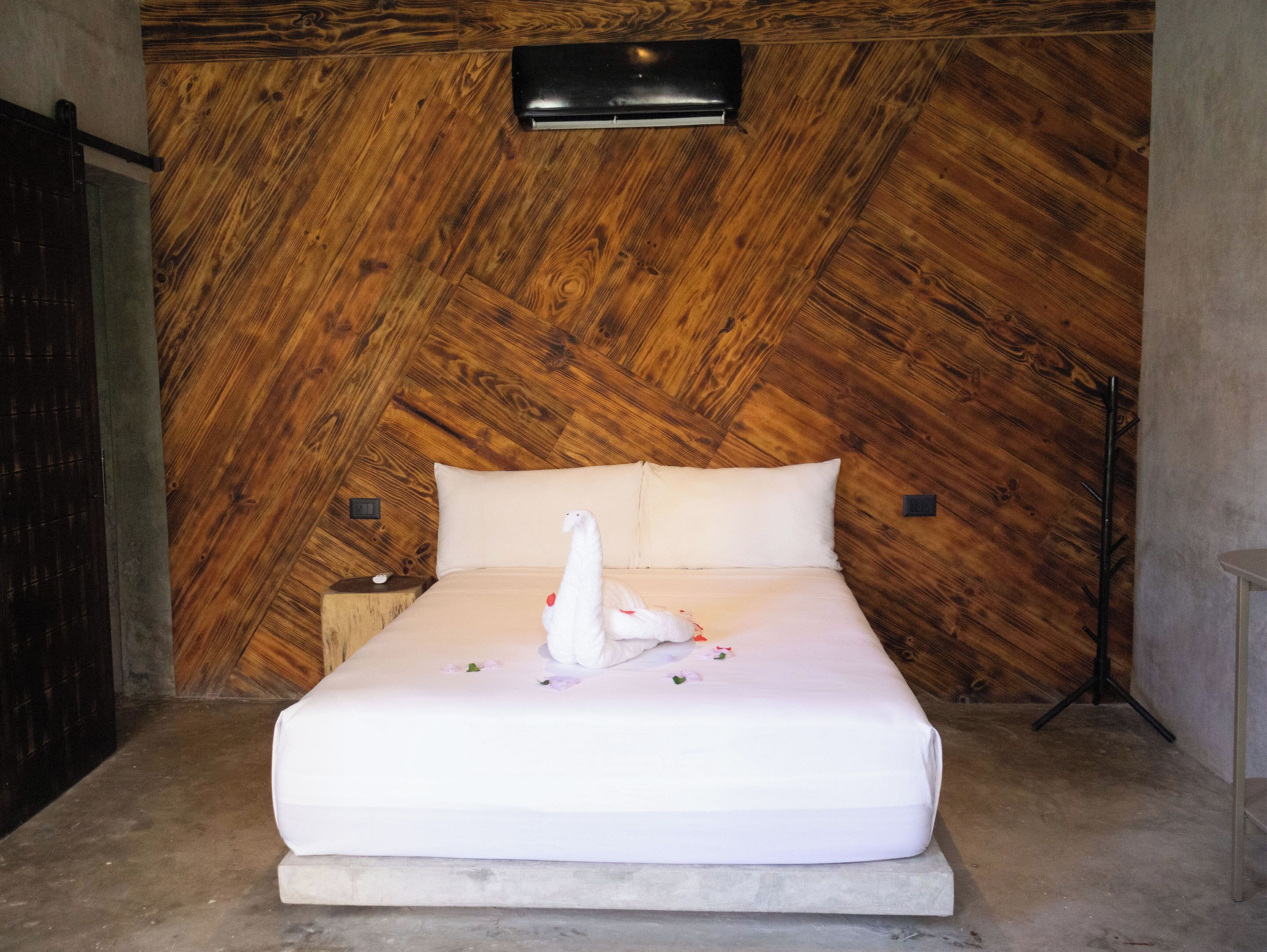 A bed with white linens, a white towel, and a decorative swan in a room with wooden paneling and a wall-mounted air conditioning unit.