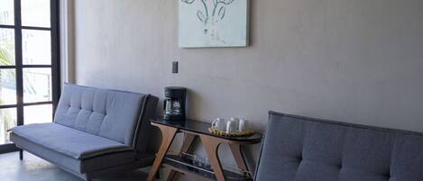 Um quarto com dois sofás cinzas, uma máquina de café e uma pequena mesa com chávenas e um tabuleiro.