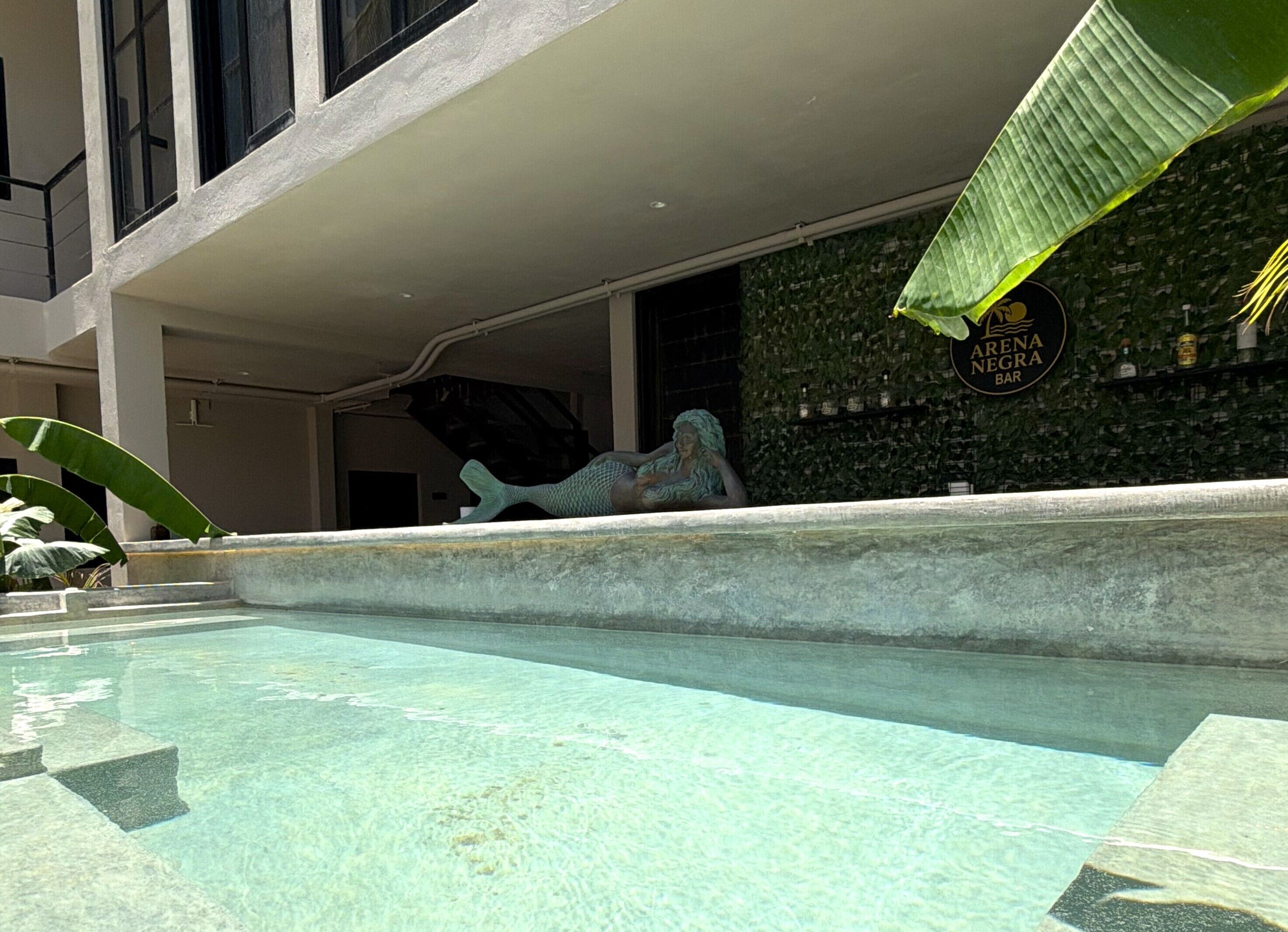 Una piscina con una estatua de una sirena, un letrero de bar y un edificio con un muro cubierto de follaje verde.