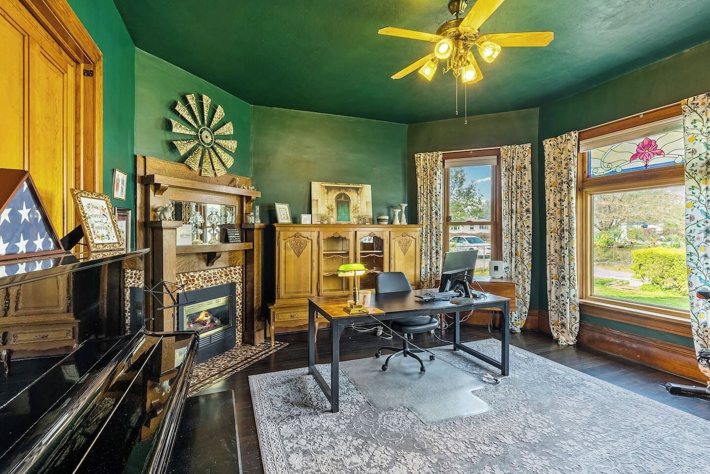 A room with a fireplace, a desk with a computer, a ceiling fan, and a view of the outdoors.