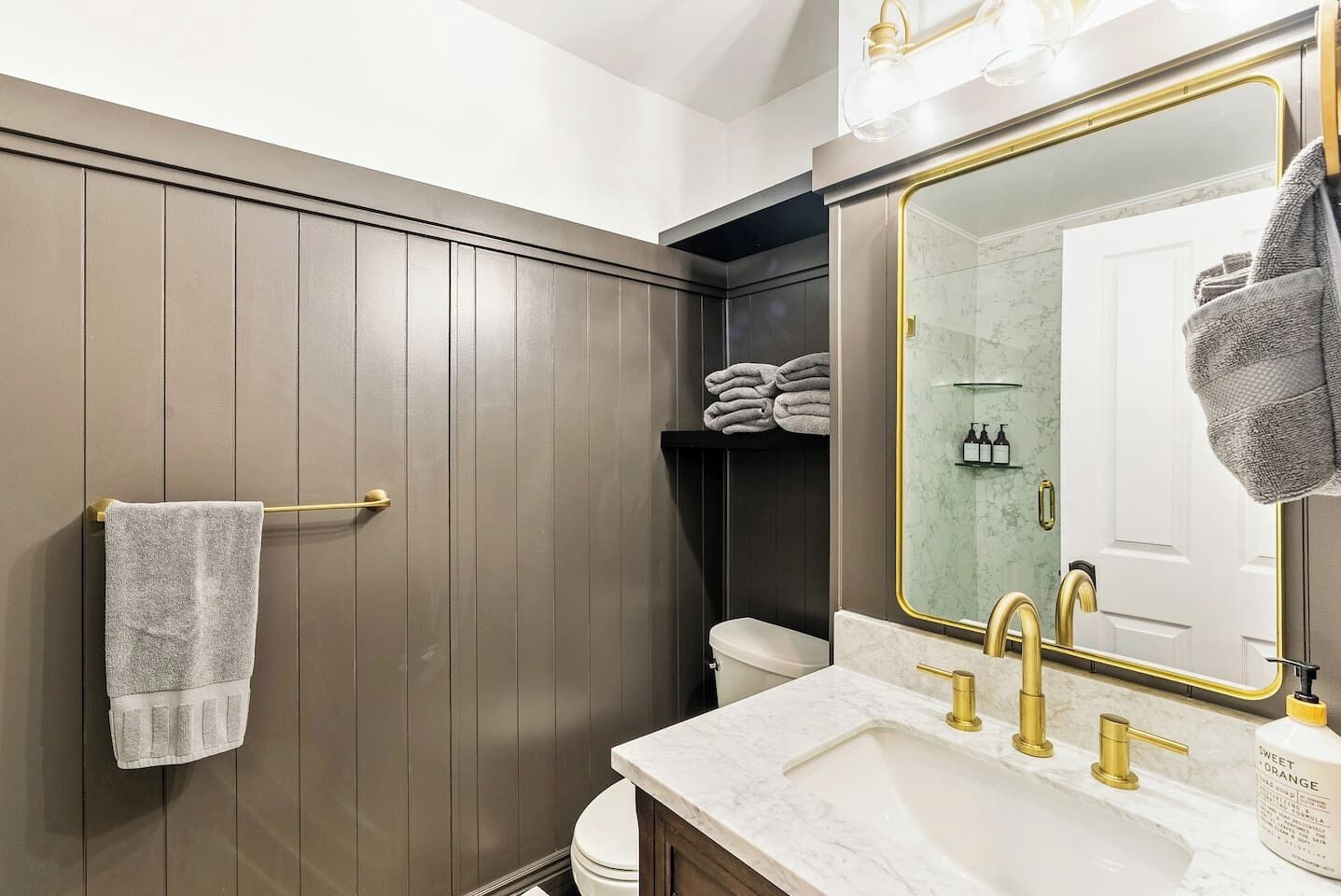 Een badkamer met een wand van houten panelen, een wit wastafelmeubel met gouden details, en een grote spiegel met een gouden lijst.