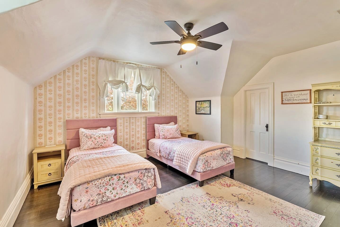 Une chambre avec deux lits, un ventilateur de plafond, une fenêtre avec des rideaux, une commode et un mur orné d’une citation.
