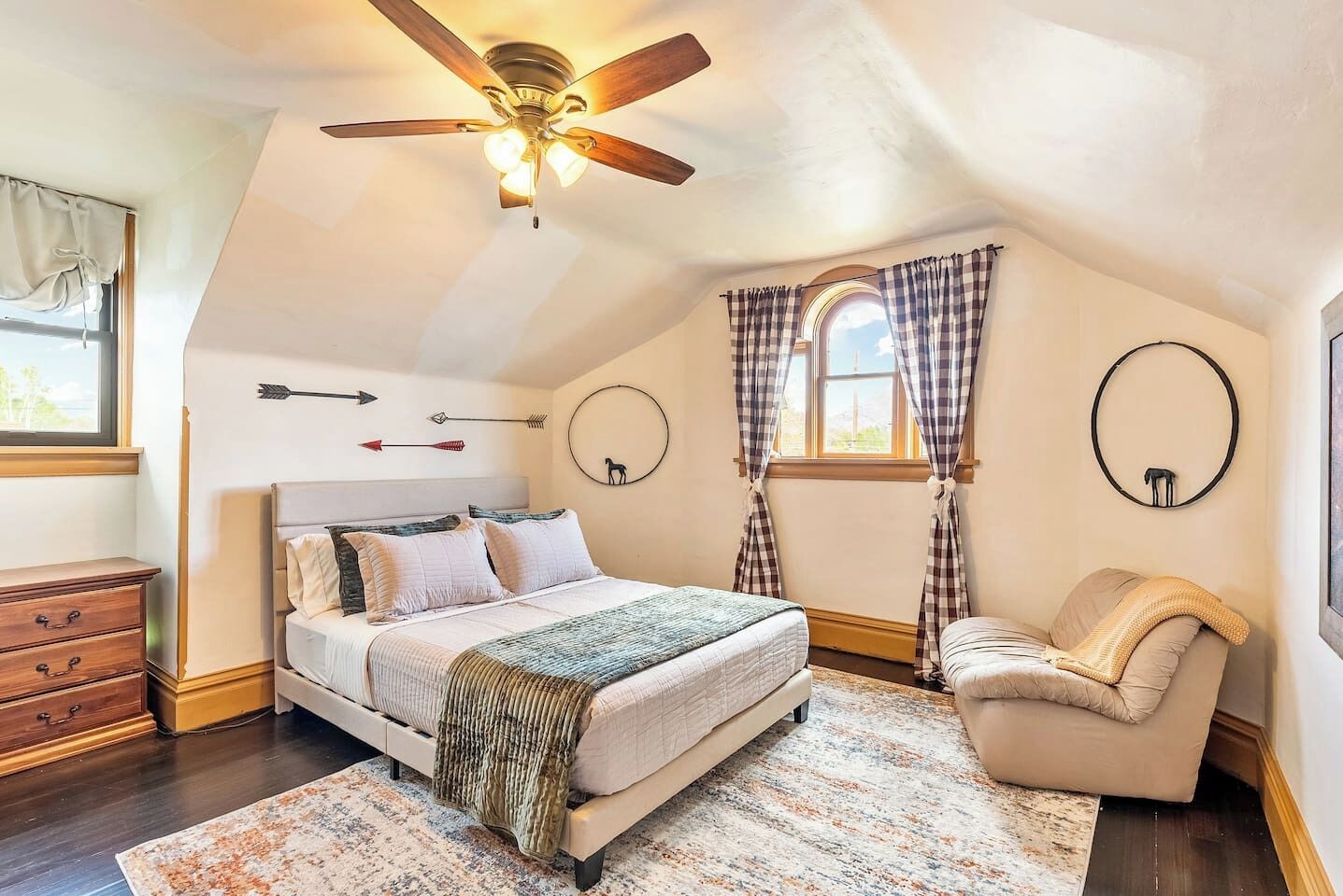 Une chambre à coucher comprenant un lit, une chaise, une commode, un ventilateur de plafond et une fenêtre avec des rideaux.