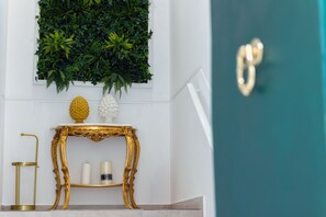 Ein Zimmer mit weißer Wand, einer grünen Tür, einem verzierten goldenen Tisch mit weißer Vase und einer Wand aus Pflanzen.