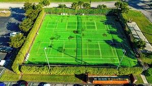 Vista aérea de uma quadra de tênis cercada por uma grade, com estacionamento e áreas verdes próximas.