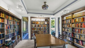 Uma biblioteca espaçosa com uma grande mesa de madeira, várias estantes e um ventilador de teto.