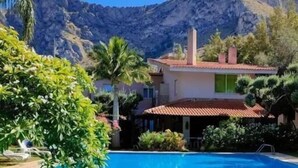 Vista de la piscina con montañas al fondo, una silla de ruedas y una casa con techo rojo.