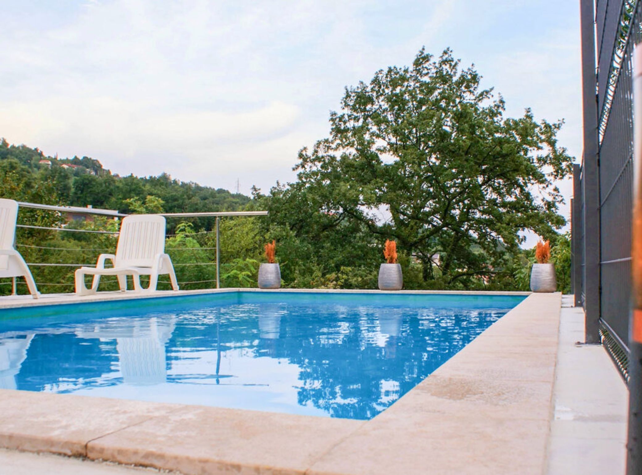 A swimming pool with white lounge chairs and potted plants, surrounded by a fence and trees.