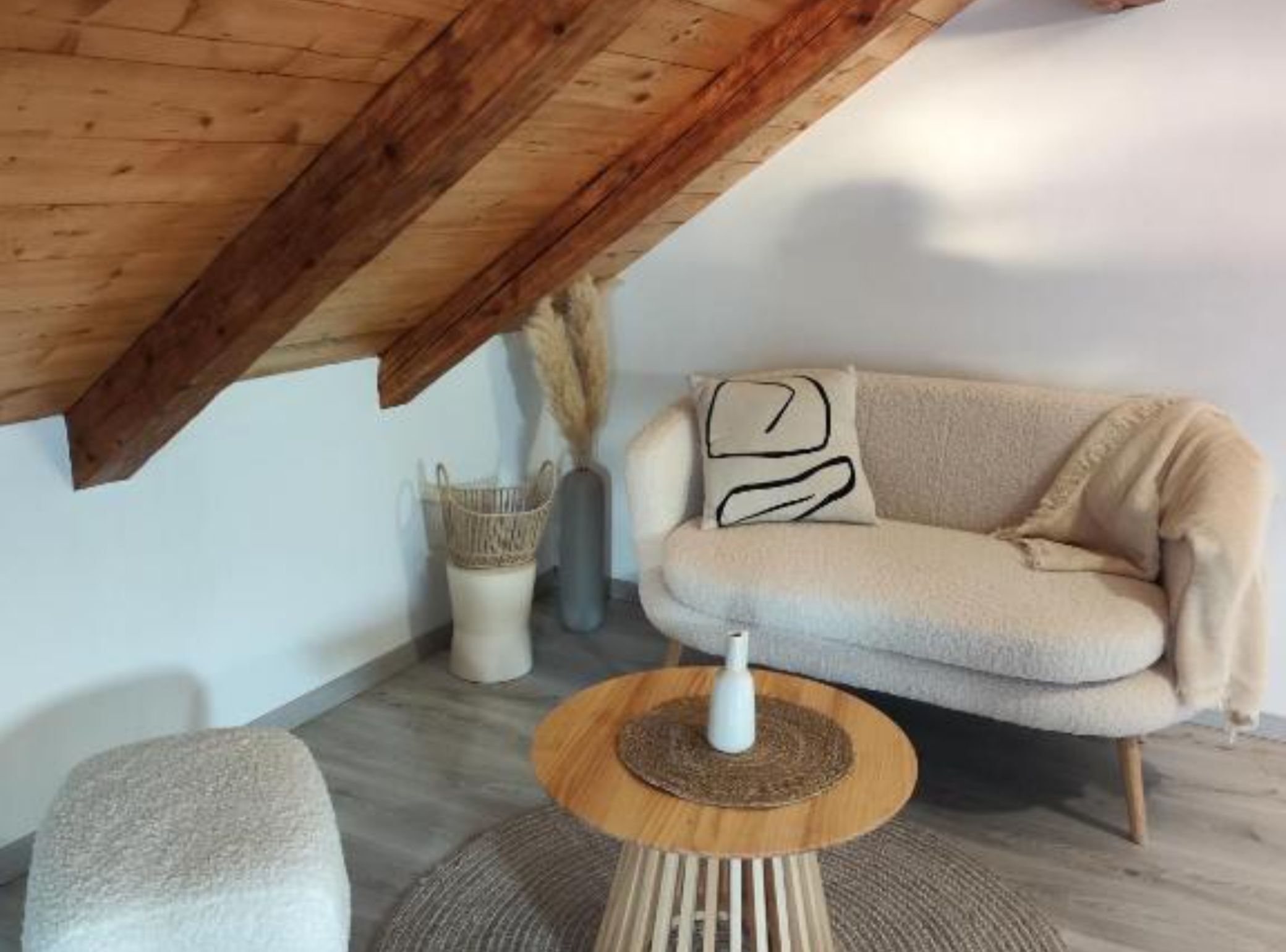 Ein gemütlicher Raum mit Holzbalkendecke, einem beigen Sofa, einem runden Holztisch und einem kleinen Hocker.