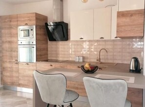 A modern kitchen with wooden cabinets, a built-in oven, a microwave, a black TV, a sink with a kettle, and two chairs. - Apartment Penthouse Ivan (Drnis)