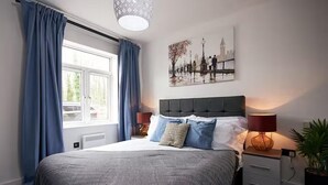 A bedroom with a bed, a window with curtains, a wall-mounted artwork, bedside lamps, and a nightstand with a plant.