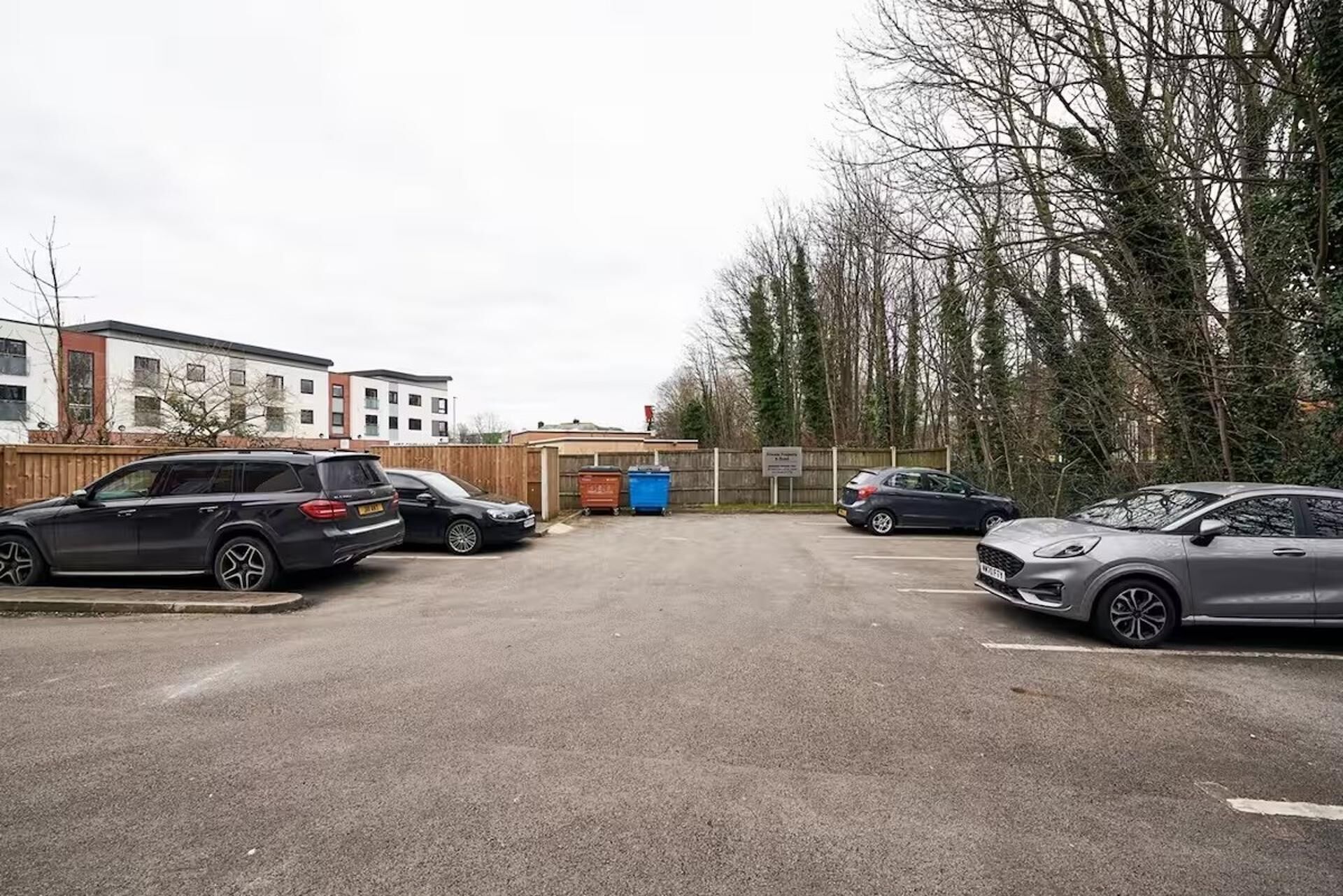 A parking lot with several cars, a building with multiple windows, and a fence with trees in the background.