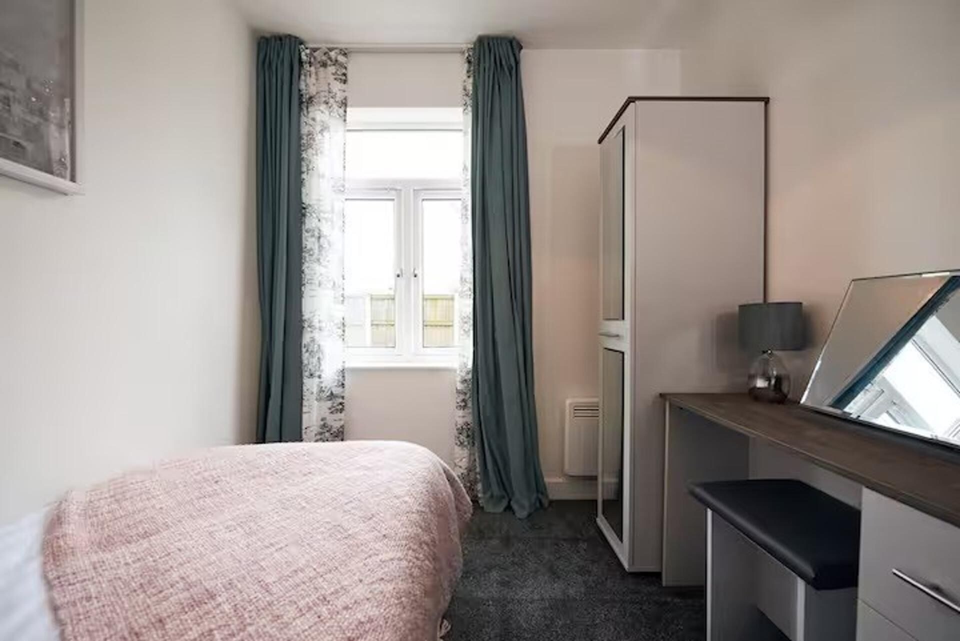 A hotel room with a bed, a desk, a mirror, a lamp, and a window with curtains.