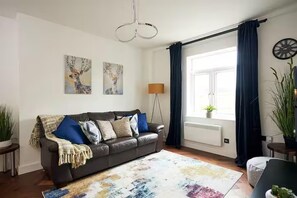 A living room with a sofa, a floor lamp, artwork, and a window with curtains.