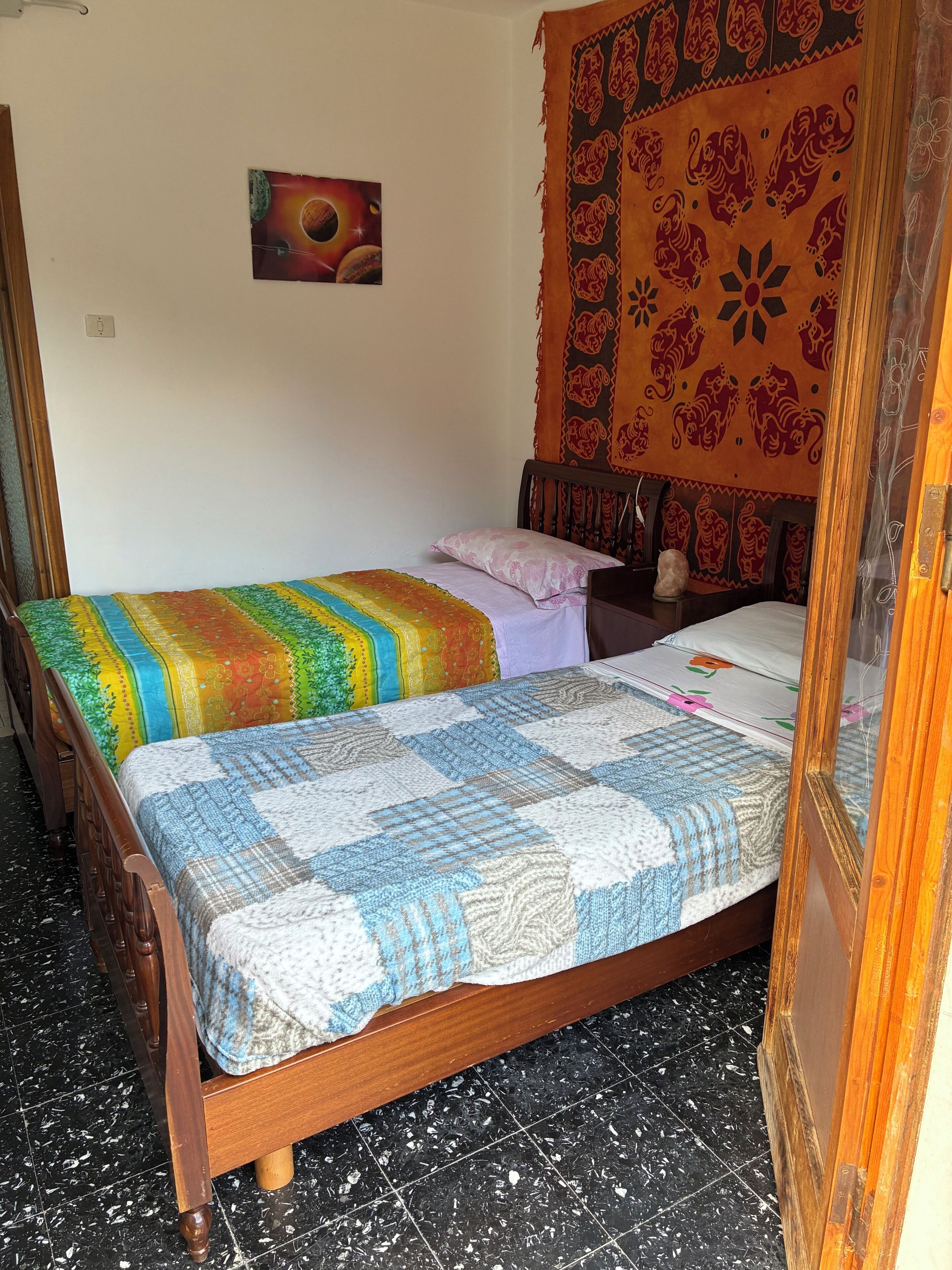 Quarto com cama de madeira, colcha colorida, cabeceira com tapeçaria e um quadro fixado na parede.