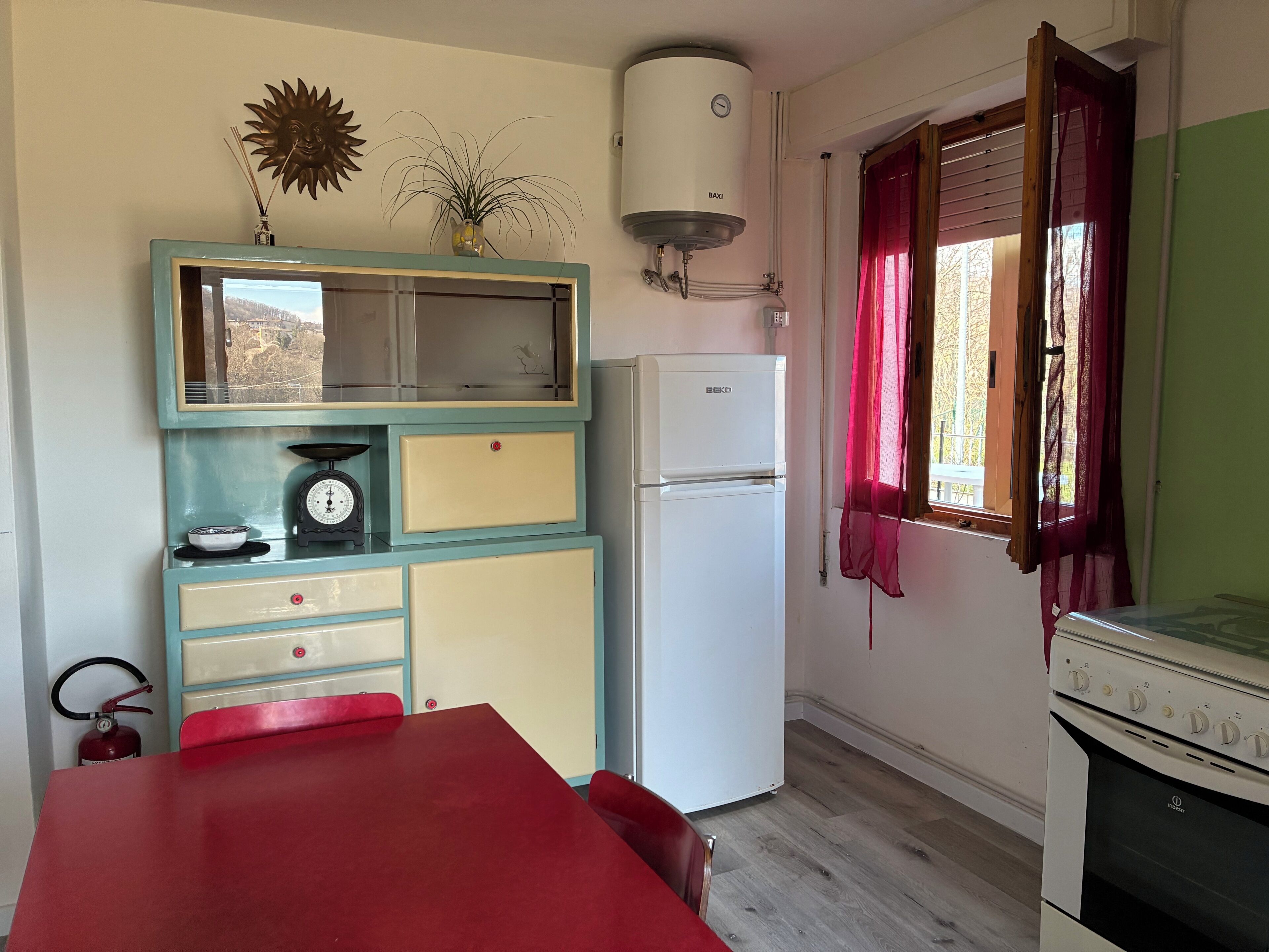 Eine Küche mit einem alten Herd, einem Kühlschrank, einer Waage und einem Fenster mit Vorhängen.