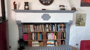 A fireplace with a mantel decorated with a crest and a plaque, a bookshelf filled with books, a red chair, and a wall-mounted painting. - Apartment in the midst of nature that will help regenerate your body and mind (Castiglione dei Pepoli)