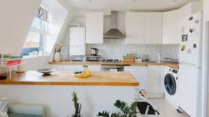 Une cuisine moderne avec des meubles blancs, un plan de travail en bois, une cuisinière à gaz, un four encastré, un lave-linge et une fenêtre qui laisse entrer la lumière naturelle.