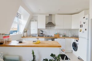 A modern kitchen with white cabinetry, a wooden countertop, a gas stove, a built-in oven, a washing machine, and a window providing natural light. - Sunny 1BD Flat in the Heart of Maida Vale (Greater London)