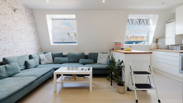 Un salon moderne avec un canapé d’angle, une table basse blanche et un coin cuisine doté d’un plan de travail et de meubles de rangement blancs.