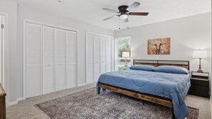 Une chambre à coucher comprenant un lit, un ventilateur de plafond, une table de chevet, une lampe et une fenêtre avec des stores.