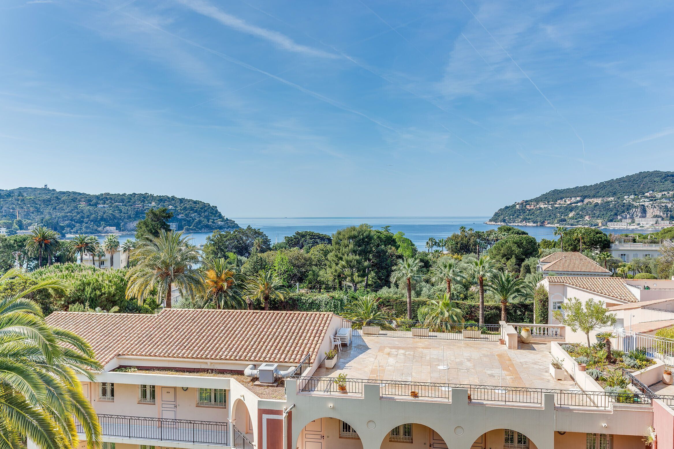 A coastal residential area with terracotta-roofed buildings, a clear blue sky, and a view of the sea.