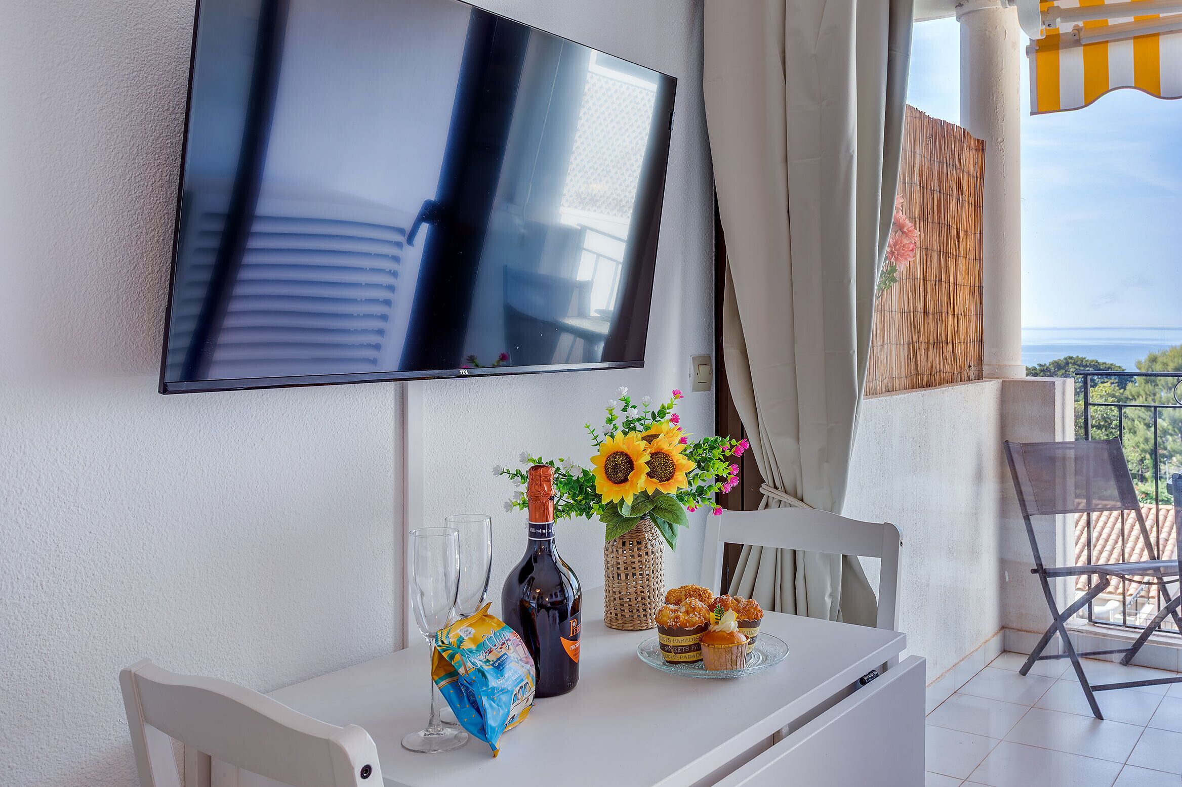 Une pièce avec un téléviseur fixé au mur, une table avec une bouteille, des verres, un vase avec des fleurs et une chaise.