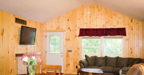 A wooden-paneled room with a television mounted on the wall, a fireplace, a couch, and a window with a view of greenery. - Scotts Creek Cabins - Hillside (Logan)