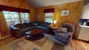Ein gemütliches Wohnzimmer mit einem Ecksofa, einem kleinen Couchtisch, einem Sessel und einer Lampe. Zwei Fenster mit Vorhängen, ein gerahmtes Bild an der Wand und eine Küchenzeile mit Schränken.