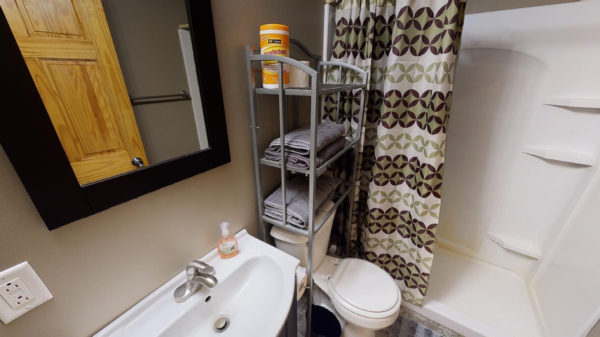 A bathroom with a white sink, toilet, and bathtub. There is a shelf with folded towels and a container of disinfectant.