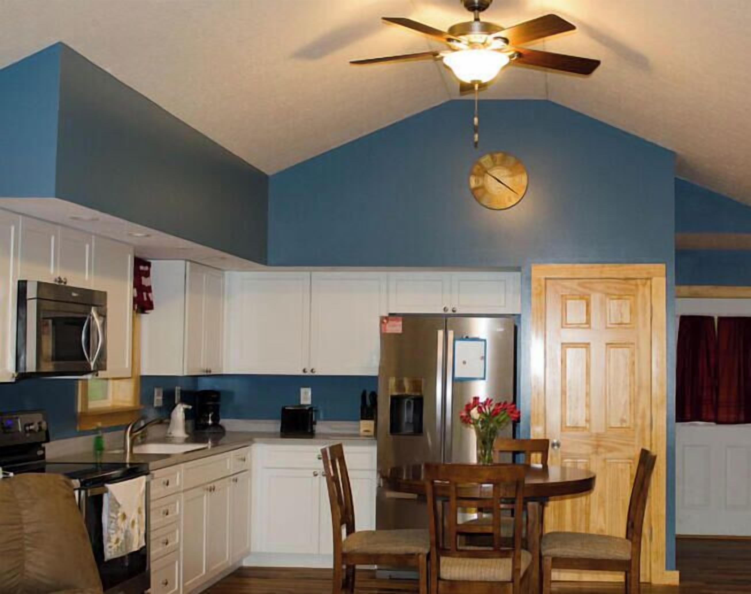 A kitchen with white cabinets, a dining table with chairs, a ceiling fan, and a clock.