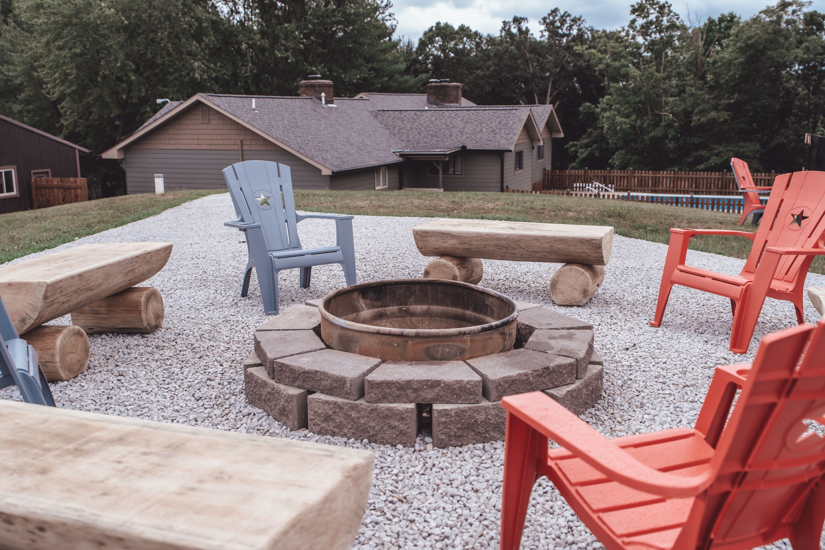 A fire pit surrounded by wooden benches and Adirondack chairs in a gravel area with a house in the background.
