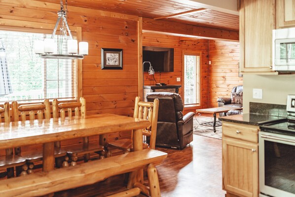 Interior de una cabaña de madera con una mesa de comedor, sillas, chimenea, televisor y zona de cocina.