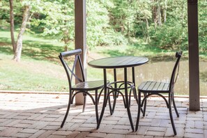 A covered patio with a round table and two chairs, surrounded by a brick floor and a view of a pond. - Scotts Creek Cabins - Hem Loch (Logan)