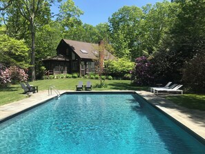 A rectangular swimming pool with clear water, surrounded by a paved deck. There are lounge chairs on the deck, and a wooden house with a sloped roof is visible in the background. - Perfect 3-bedroom house with pool on lush treed garden flag lot  in East Hampton (East Hampton)