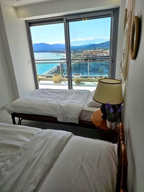 Ein Hotelzimmer mit zwei Betten, einem großen Fenster mit Blick auf das Meer und Gebäude sowie einer Lampe auf einem Nachttisch.