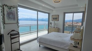 Ein Schlafzimmer mit einem Bett, einem Nachttisch mit Lampe, einem Sessel, einer Kommode und einem großen Fenster mit Blick auf das Meer und die Berge.