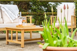 A wooden table with a bottle and glass, a tray, and a bowl of snacks, set on a patio with a sofa and chairs. - The Cottage at Tuffon Hall, Sible Hedingham, Suffolk (Sible Hedingham)