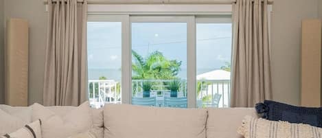 A room with a sofa and two windows with curtains, a view of the outdoors, and a balcony with plants.